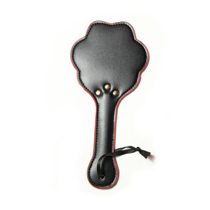 a black leather paddle with a unique design. The paddle is shaped like a paw print and features red stitching around the edges. It has three metal rivets at the base of the paw print design and a wrist strap attached to the handle for better grip and control. This type of paddle is typically used in BDSM practices for impact play, providing both a visual and tactile element to the experience.