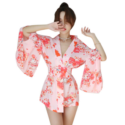 A woman is wearing a light pink floral kimono with wide, flowing sleeves and a matching belt. Her pose is casual, with one hand raised and covering part of her face. The kimono features a vibrant floral pattern in shades of red and white, adding a touch of elegance to her relaxed look.