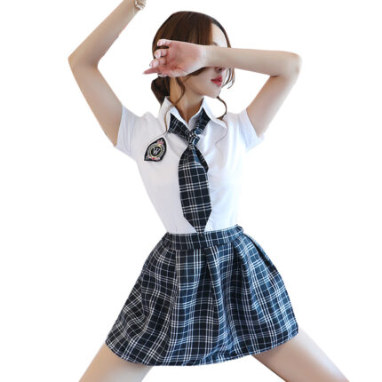 A woman is wearing a short-sleeved white shirt with a black and white plaid skirt and matching tie, styled in a school uniform fashion. Her pose is dynamic, with her arms up and crossed over her face. The outfit includes a patch on the shirt, adding to the uniform look, and her hair is loosely tied back.